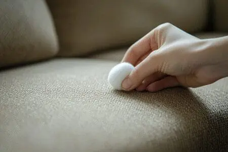 A close-up of a hand using a cotton ball to lift a pen ink stain from a sofa, showing a DIY save