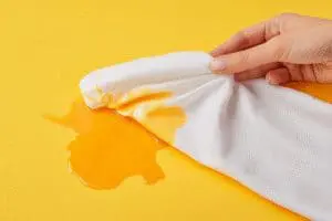 A hand dabbing a cloth to remove an orange self-tanner stain from a white shirt, showing an easy fix