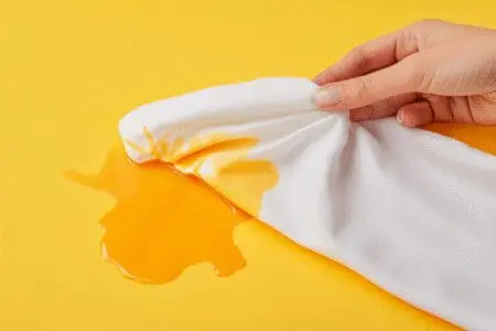 A hand dabbing a cloth to remove an orange self-tanner stain from a white shirt, showing an easy fix