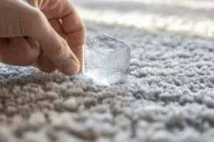 A hand using an ice cube to freeze chewing gum on a carpet for easy, damage-free removal.